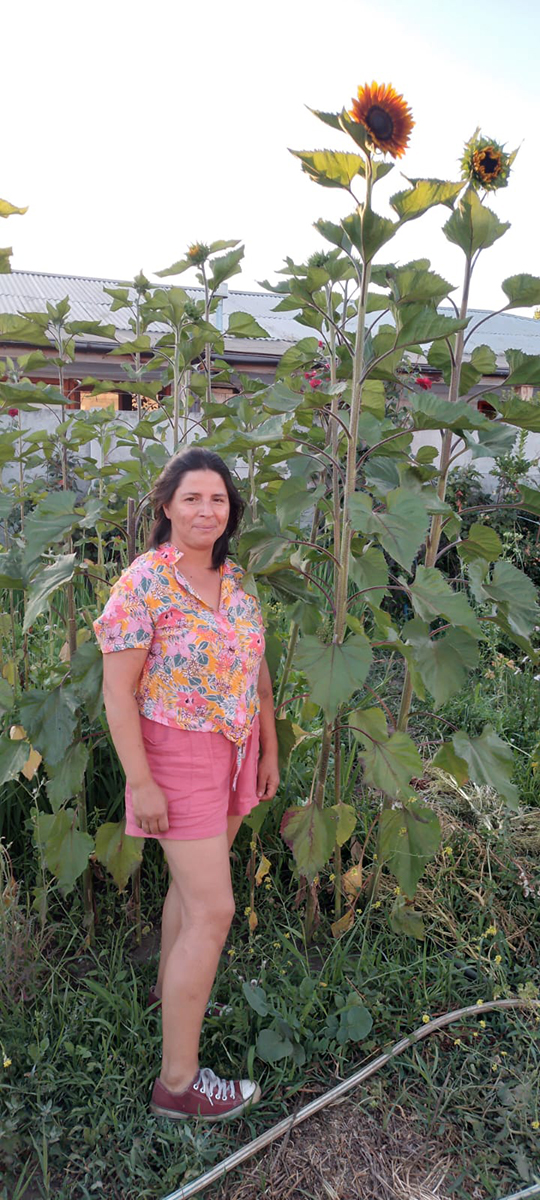 Mujer de pie en un huerto, rodeada de plantas de girasol de gran altura, vistiendo blusa floral de colores y short rosado, en un entorno rural al aire libre con vegetación abundante.