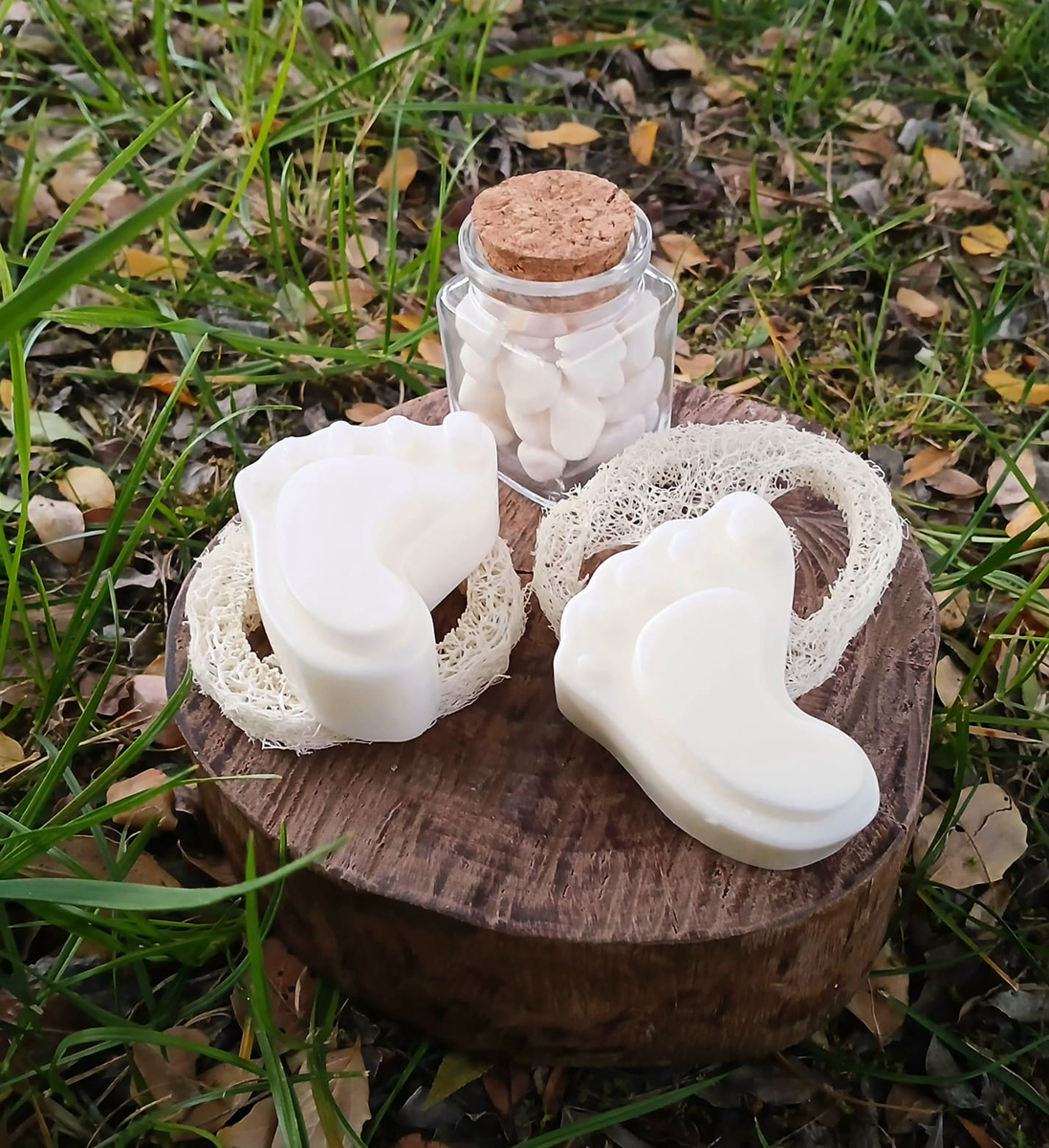 Conjunto de jabones artesanales blancos con forma orgánica dispuestos sobre una base de madera en exterior, acompañados de una esponja vegetal natural y un frasco de vidrio con tapa de corcho lleno de pequeños jabones, rodeados de pasto y hojas secas.