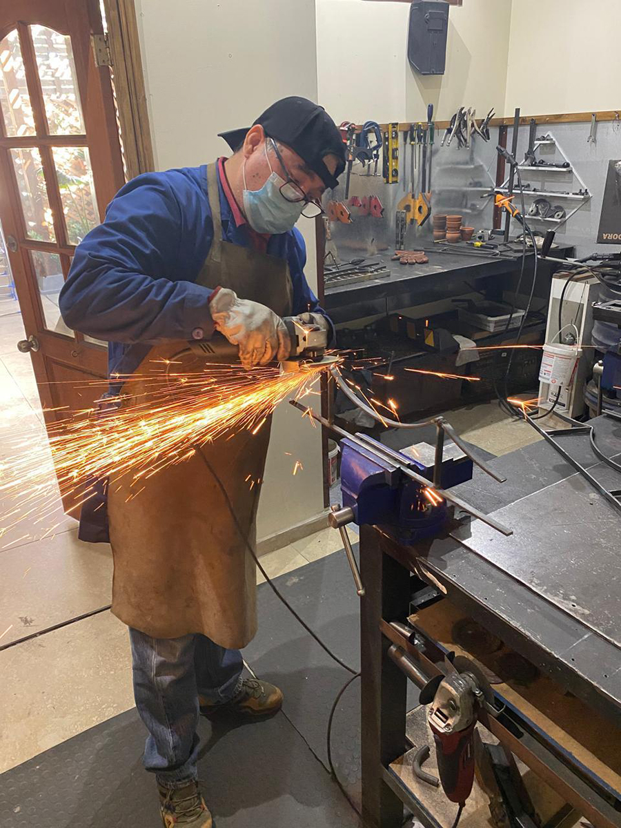 Artesano trabajando metal en un taller, usando una esmeriladora que genera chispas, con herramientas y banco de trabajo al fondo.