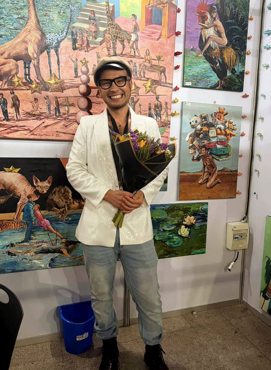 Artista sonriente sostiene un ramo de flores frente a una pared llena de pinturas coloridas de estilo surrealista y narrativo, en un espacio de exhibición artística.
