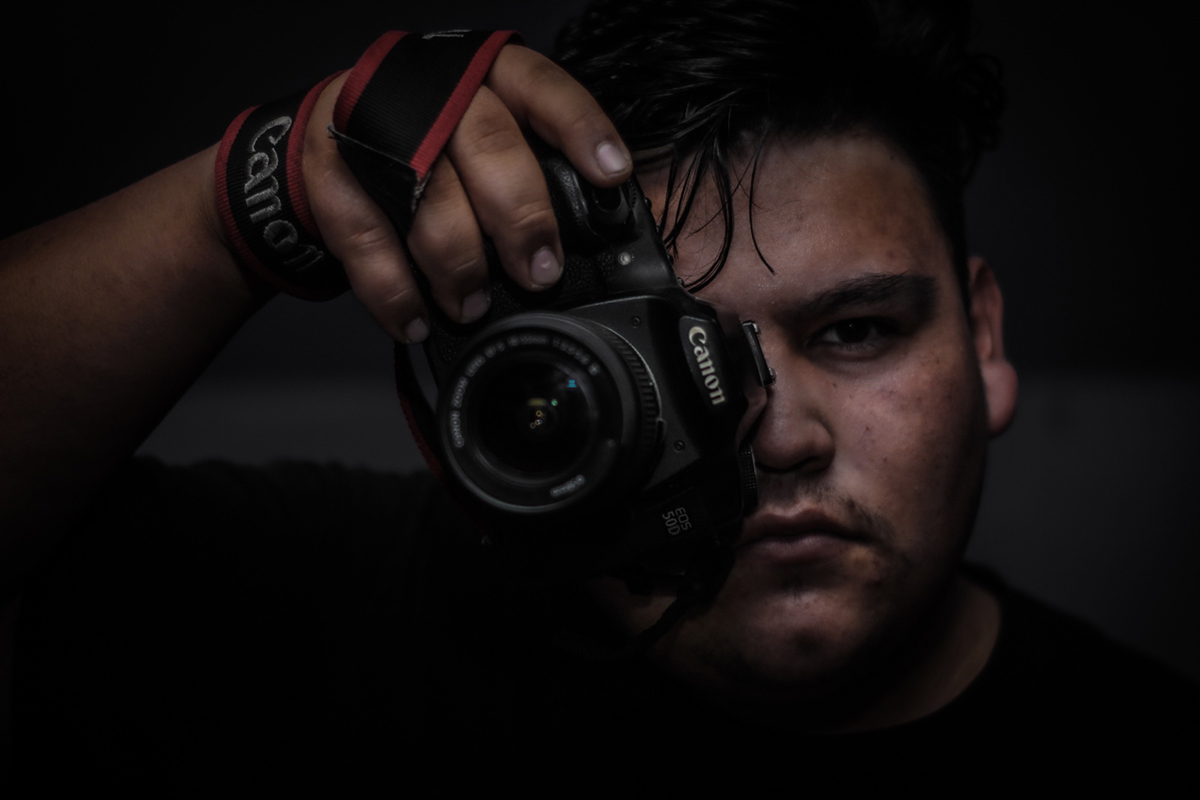 Retrato en primer plano de un fotógrafo sosteniendo una cámara réflex frente a su rostro, iluminado de forma dramática sobre un fondo oscuro.