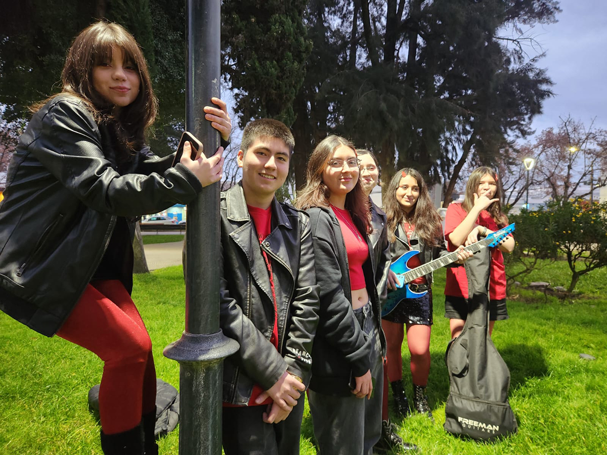 Grupo de jóvenes músicos al aire libre, de pie sobre el césped en una plaza, algunos con instrumentos y estuches, vistiendo chaquetas negras y ropa roja al atardecer.