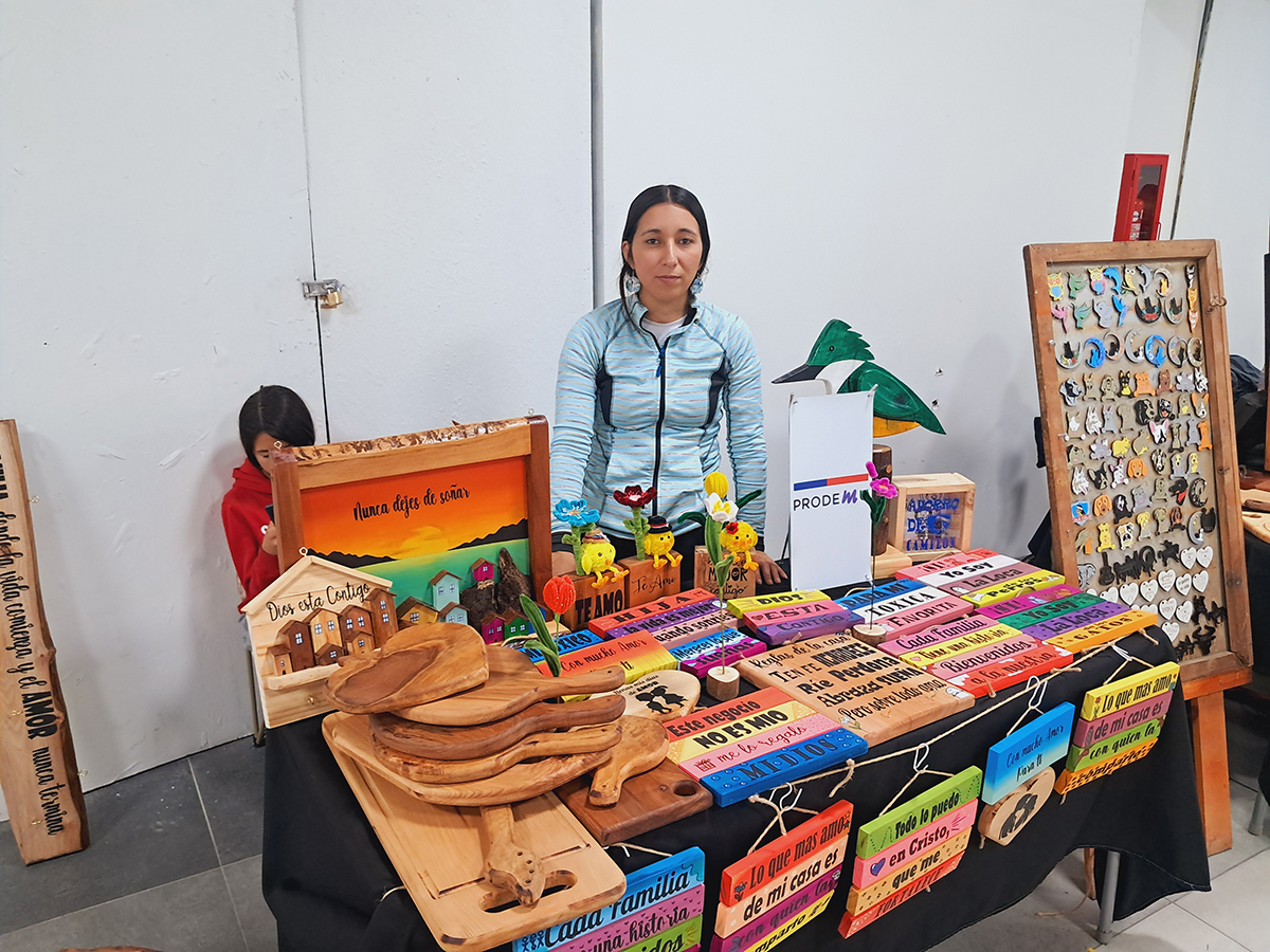 Mujer artesana de pie detrás de una mesa exhibiendo artesanía en madera, con tablas, letreros pintados y piezas decorativas coloridas, en un espacio interior de feria.
