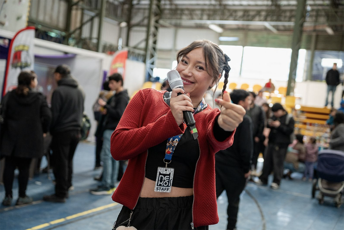 Mujer joven sosteniendo un micrófono y sonriendo, haciendo un gesto con la mano, durante un evento cultural en un gimnasio techado. Al fondo se observan personas caminando, stands y público asistente.