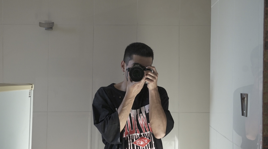 Persona frente a un espejo tomando una fotografía con cámara, rostro parcialmente cubierto por el lente, en un baño de paredes claras.