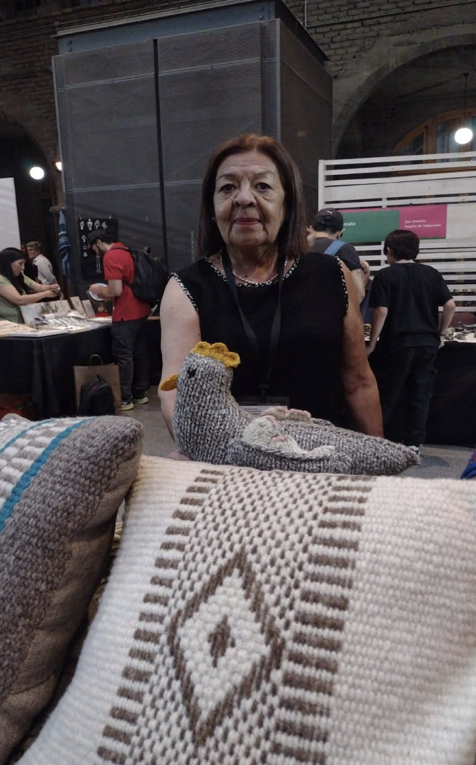 Mujer artesana presenta piezas textiles hechas a mano en un stand de la Ruta de la Lana, con gallinas tejidas, cojines y textiles de lana natural en distintos colores.