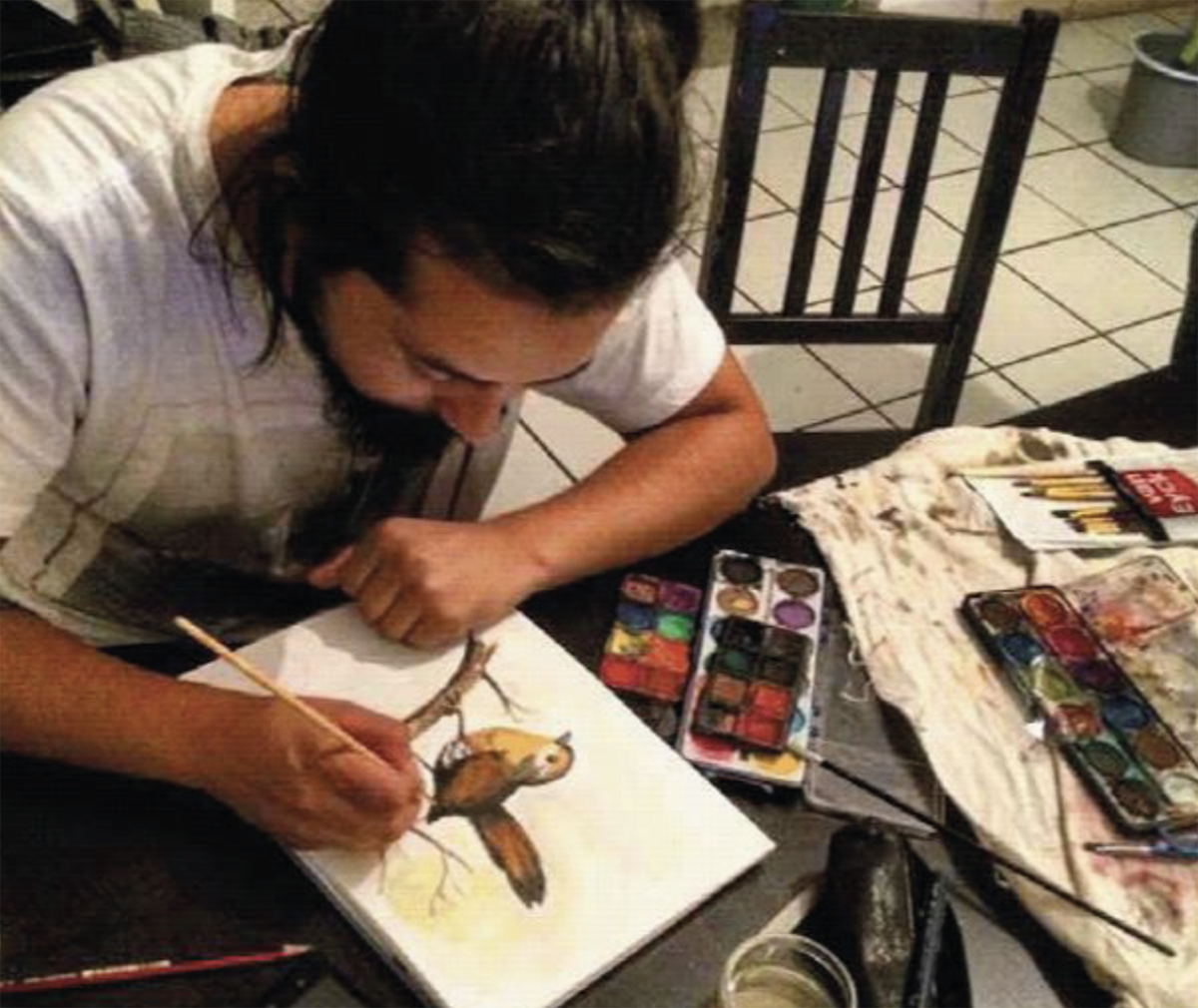 Artista trabajando en una ilustración de ave con acuarela, rodeado de pinceles, paleta de colores y materiales de pintura sobre una mesa.