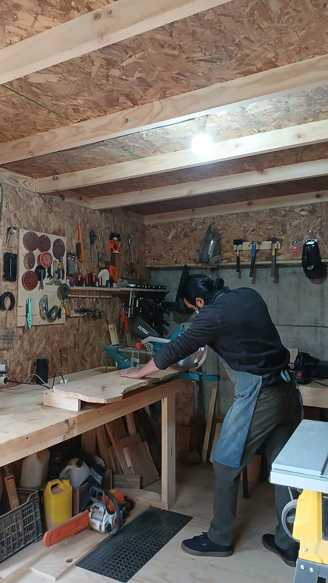 Persona trabajando madera en un taller artesanal, utilizando una sierra eléctrica sobre una mesa de trabajo rodeada de herramientas manuales y máquinas.