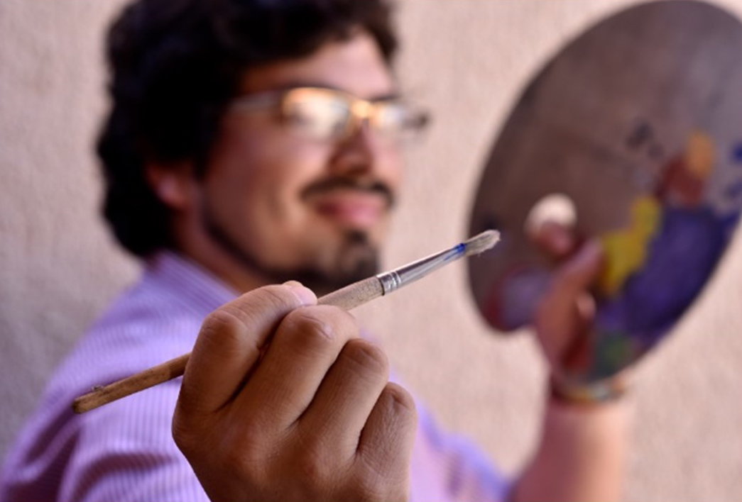 Hombre con barba y lentes sostiene un pincel frente a la cámara mientras pinta sobre una paleta, en un ambiente de taller artístico.