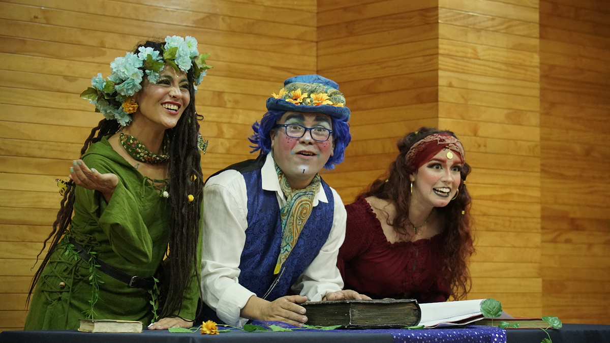 Tres intérpretes caracterizados con vestuario teatral y coronas de flores participan en una presentación escénica, apoyados sobre una mesa con un libro abierto frente al público.