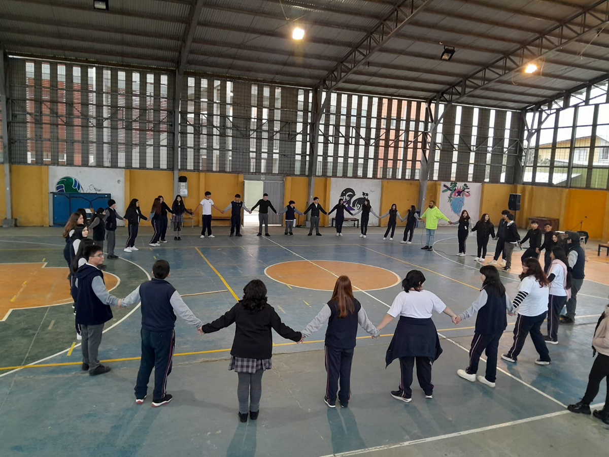Grupo de estudiantes tomados de las manos formando un gran círculo en el centro de un gimnasio techado, participando en una actividad comunitaria o educativa.