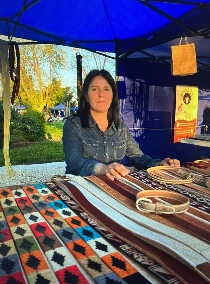 Artesana atendiendo un puesto en feria al aire libre, con una mesa cubierta de cinturones y piezas de cuero artesanal con diseños textiles de colores.