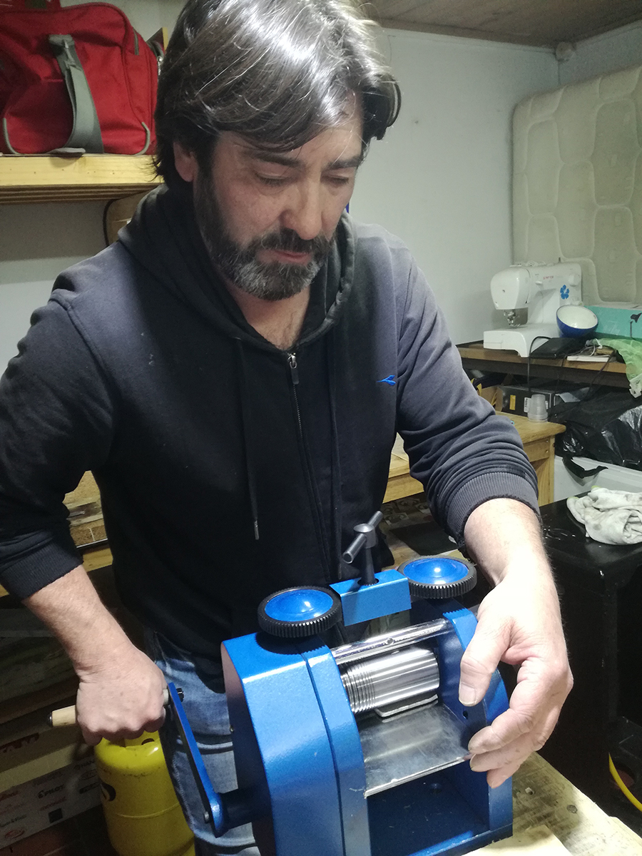 Orfebre trabajando en su taller, manipulando una laminadora manual para dar forma al metal, rodeado de herramientas y materiales de trabajo.