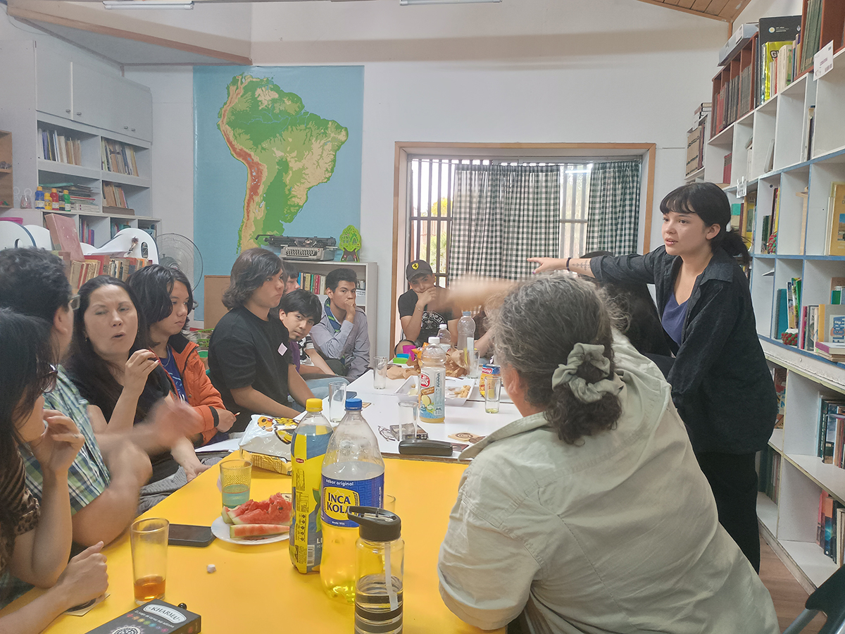 PERSONAS DIVERSAS EDADES COMPARTIENDO EN UNA MESA LARGA, BEBIDAS Y GALLETAS. ESPACIO ES UNA BIBLIOTECA PUBLICA.