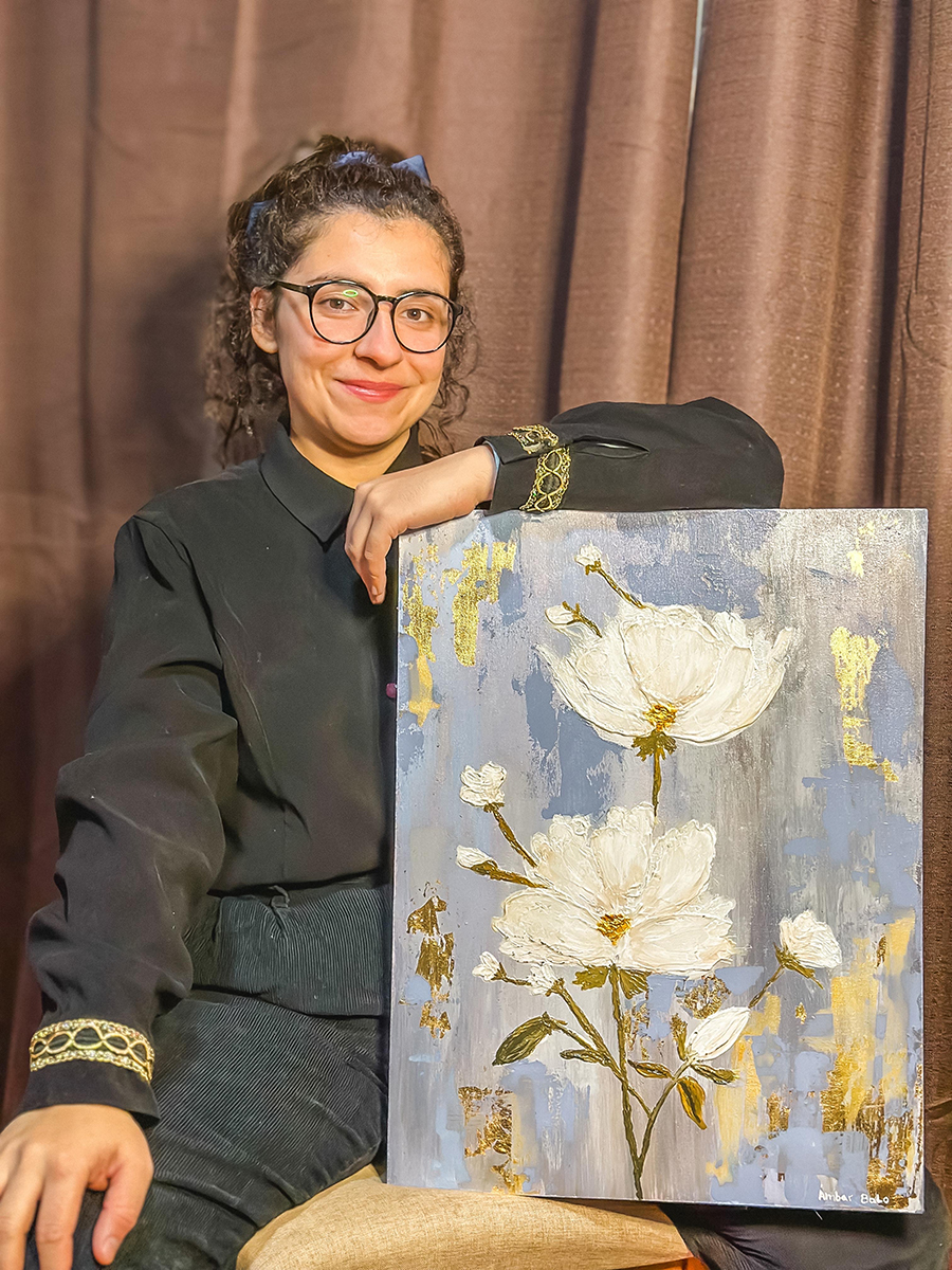 Mujer posando en interior junto a una pintura de flores blancas sobre fondo azul y gris, con detalles dorados y textura en relieve.