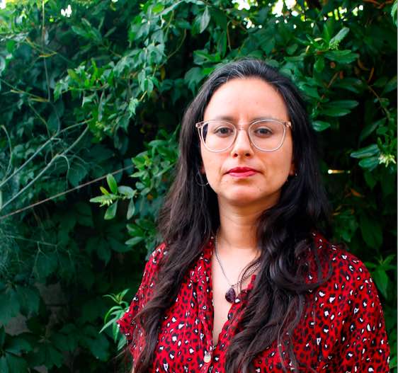 Retrato fotográfico de una mujer con anteojos, cabello largo y blusa roja con patrón, posando al aire libre frente a un fondo de vegetación verde.