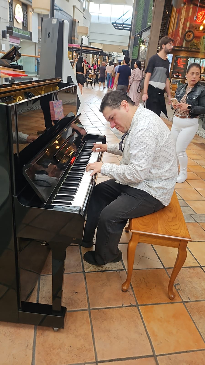 Pianista tocando un piano de cola en un espacio público tipo centro comercial, con personas caminando y observando alrededor.