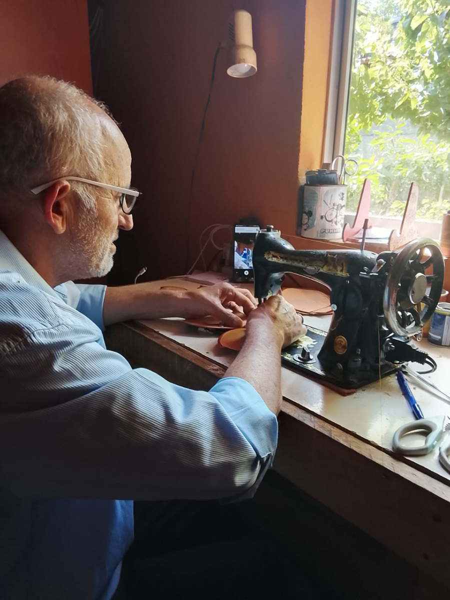 Artesano mayor trabajando cuero en una máquina de coser antigua, sentado junto a una ventana que ilumina su mesa de trabajo con herramientas y piezas en proceso.