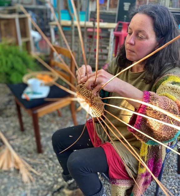 Artesana sentada al aire libre tejiendo fibras vegetales con ambas manos, rodeada de varillas largas que forman la estructura de una pieza artesanal.
