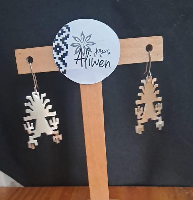 Aros de joyería mapuche en metal, con diseño geométrico tradicional, colgados en un exhibidor de madera con la marca “Joyas Afliwen”.