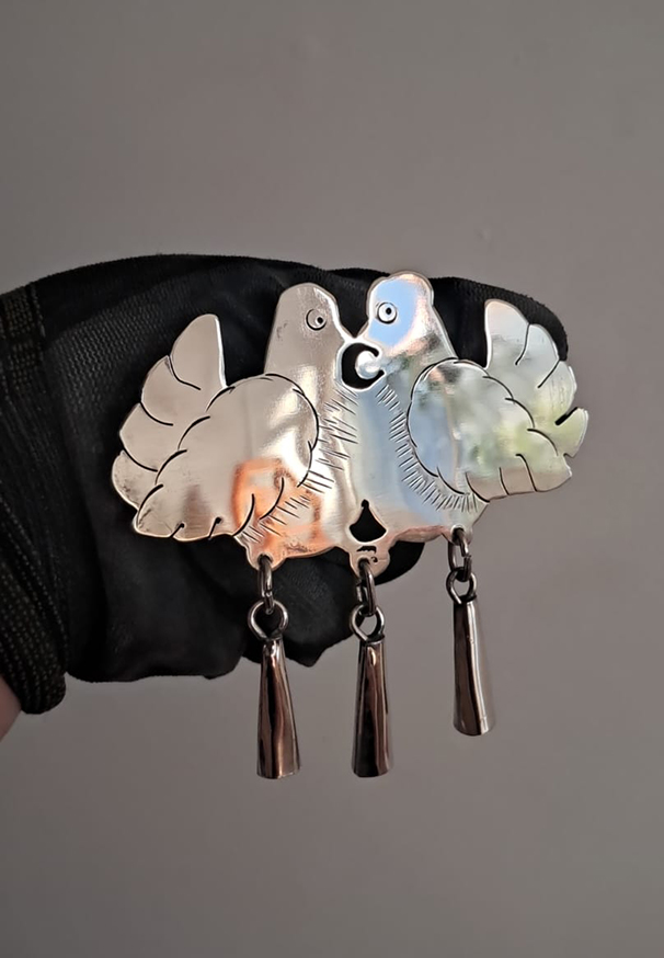 Prendedor artesanal de metal con dos aves enfrentadas y alas extendidas, con pequeñas piezas colgantes, sostenido sobre una mano con guante negro y fondo neutro.