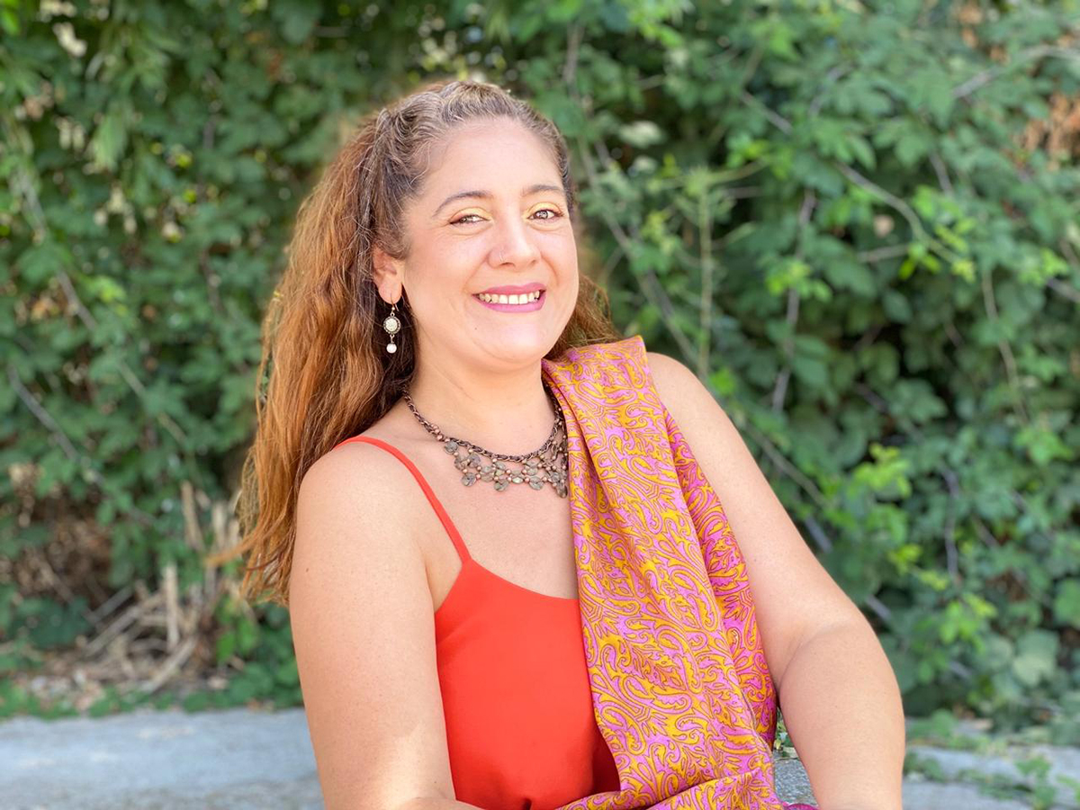 Mujer adulta sonriente al aire libre, con cabello recogido, vestida con top rojo y chal estampado en tonos rosados y dorados. Está frente a un fondo de vegetación verde, transmitiendo cercanía y alegría.