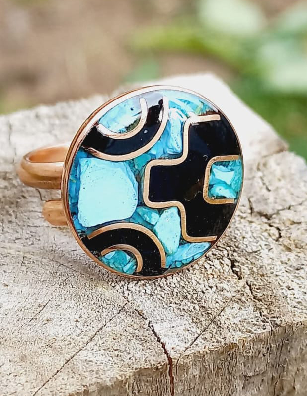 Anillo circular artesanal sobre tronco de madera, elaborado en metal cobrizo con diseño abstracto en negro y turquesa, con incrustaciones de piedra y líneas curvas decorativas.