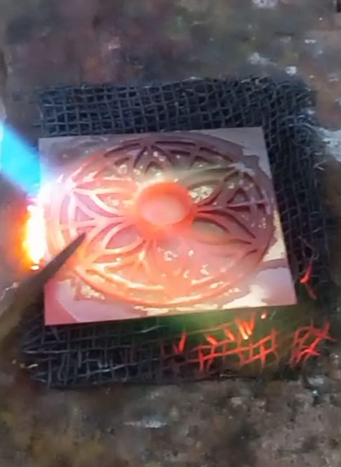 Pieza circular de metal siendo trabajada con soplete, mostrando el proceso artesanal de soldadura o fundición sobre una rejilla metálica.