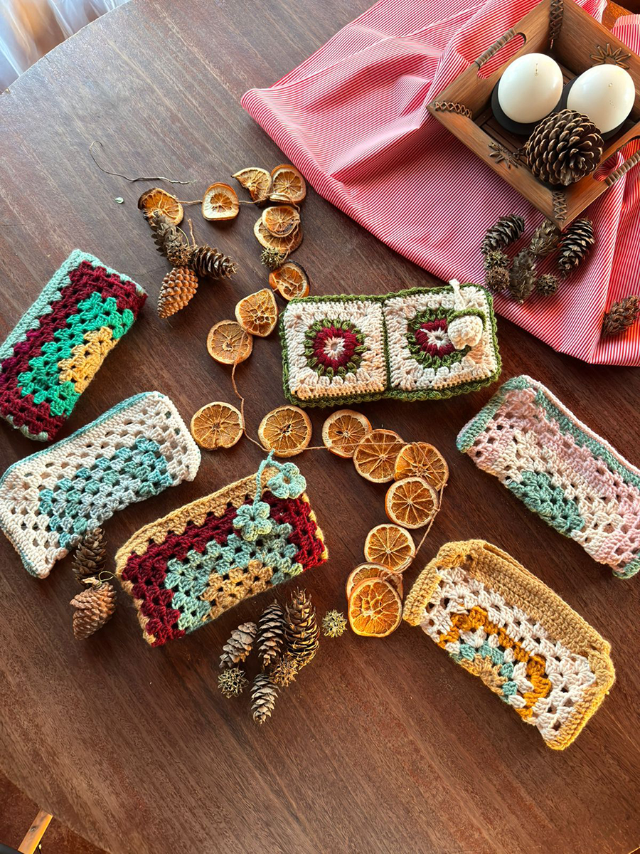 Billeteras y estuches tejidos a crochet en colores naturales y tonos navideños, dispuestos de forma circular sobre una mesa de madera junto a piñas y rodajas de naranja seca.