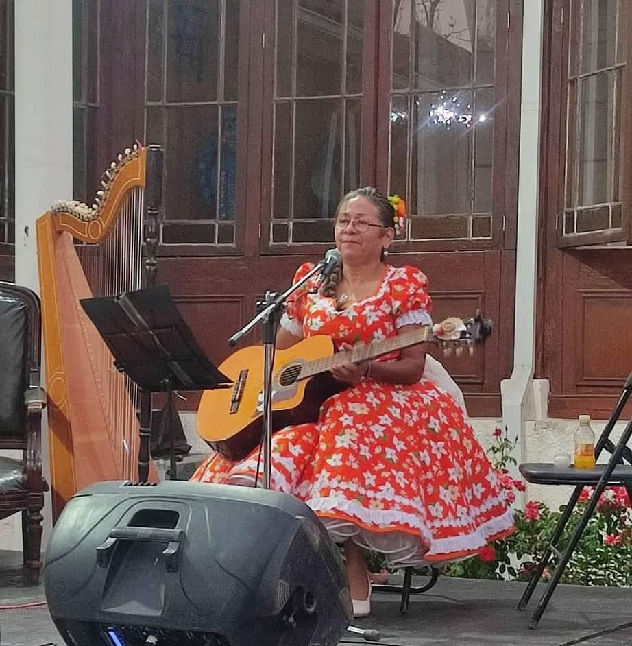 Mujer sentada en un escenario interior, vestida con traje tradicional rojo con flores blancas, tocando guitarra y cantando frente a un micrófono; a su lado se observa un arpa y un atril con partituras.