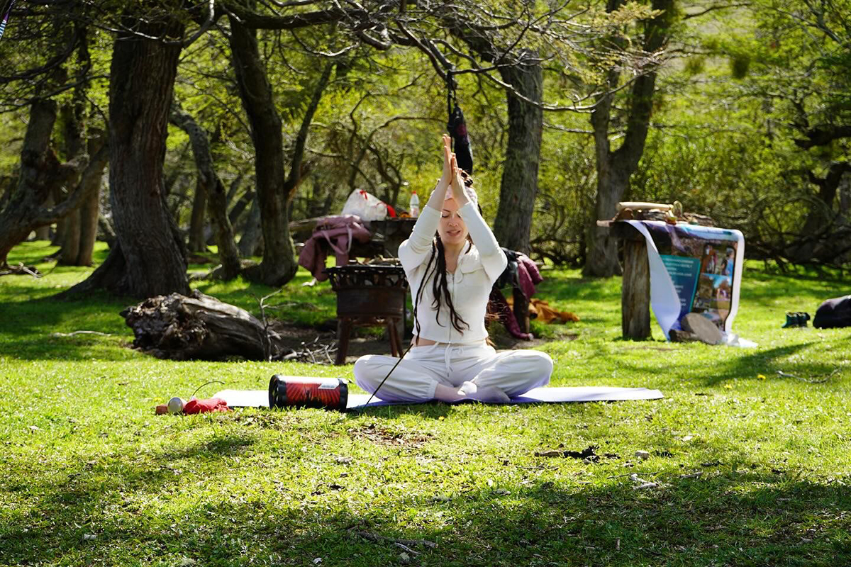 Mujer sentada en posición de meditación sobre una esterilla en un bosque, con las manos juntas frente al pecho, rodeada de árboles y luz natural.