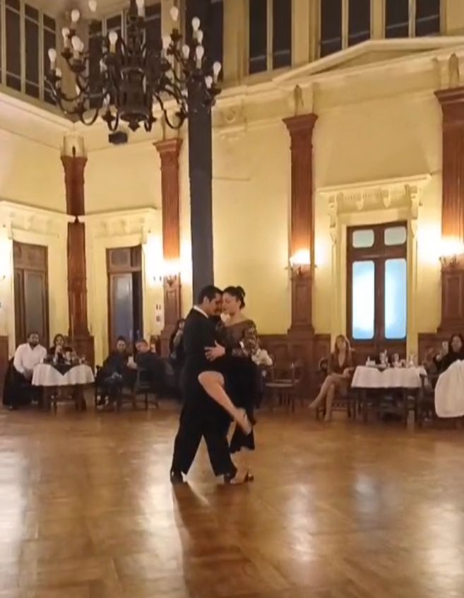Pareja interpretando tango en una milonga dentro de un salón elegante, rodeados de público sentado en mesas.