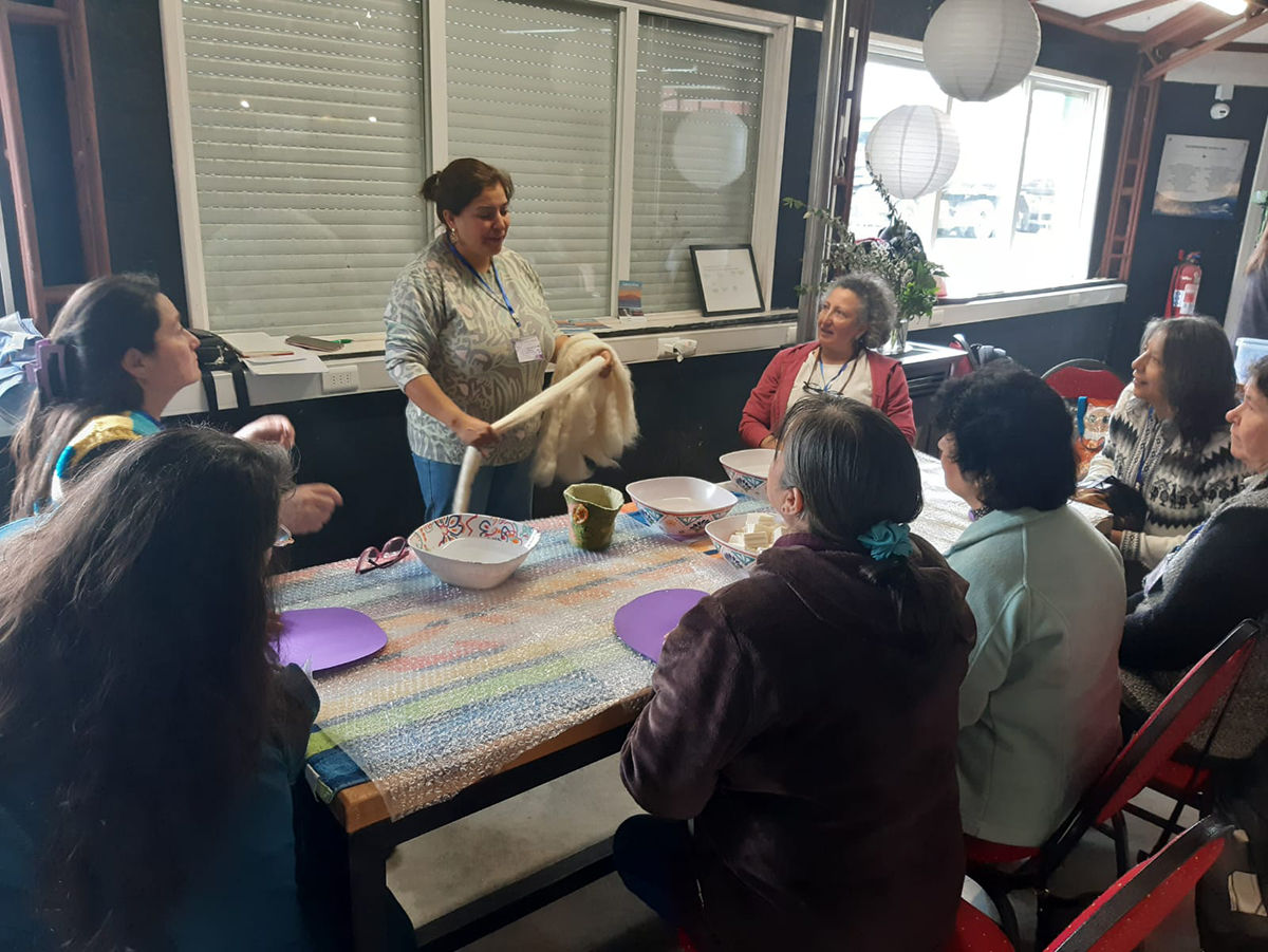 Taller comunitario de artesanía textil, donde una mujer explica el trabajo con lana a un grupo de participantes sentadas alrededor de una mesa, en un espacio interior iluminado y colaborativo.