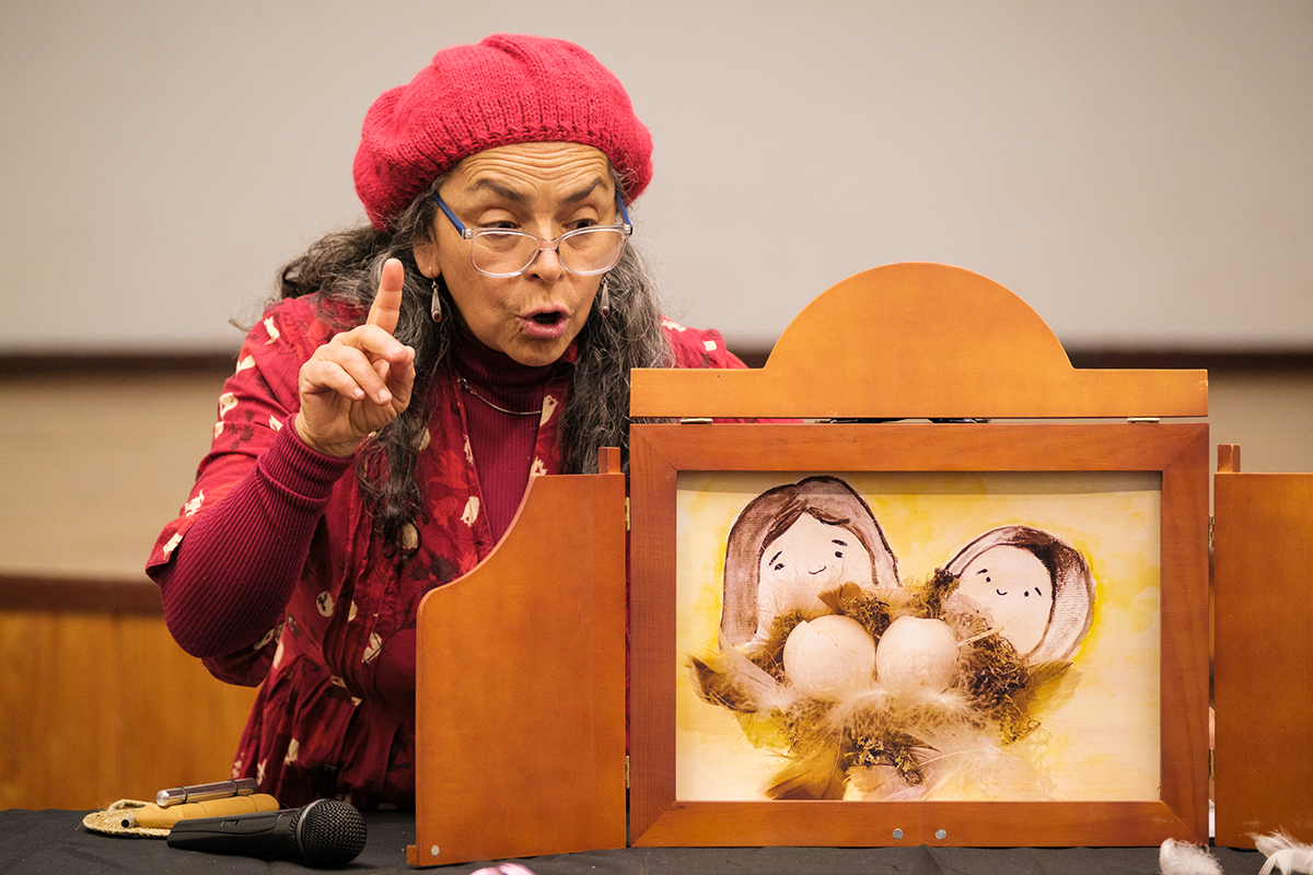 Mujer mayor narrando un cuento con un teatrillo de madera, señalando con el dedo una escena ilustrada con personajes y huevos, durante una actividad cultural en interior.