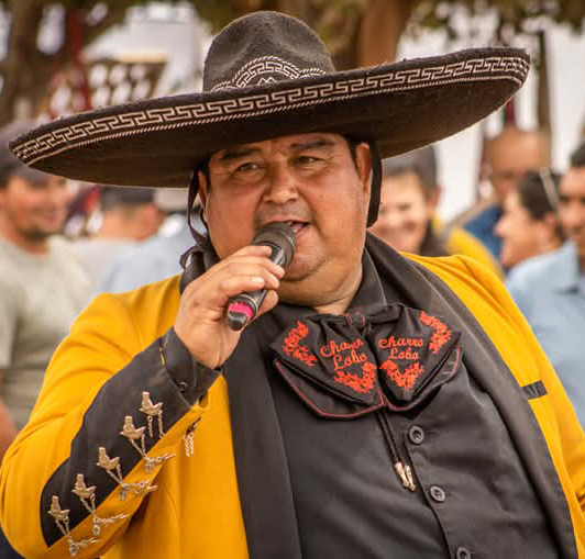 Cantante con sombrero charro y chaqueta amarilla, actuando al aire libre frente a un público, sosteniendo un micrófono mientras canta.