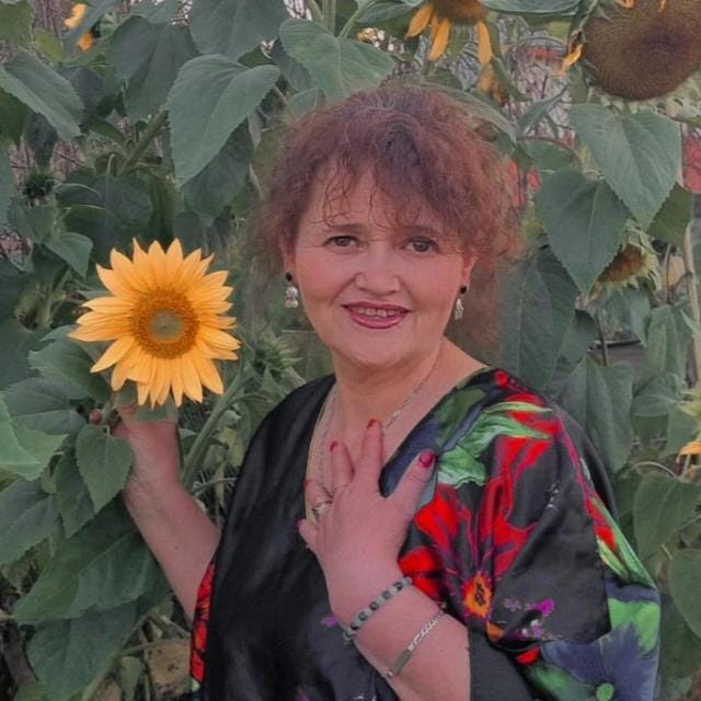 MUJER SONRIENDO, PELO CORTO,BLUSA COLORIDA, PARADA DELANTE DE GIRASOLES. SOSTIENE UNA FLOR EN SU MANO DERECHA