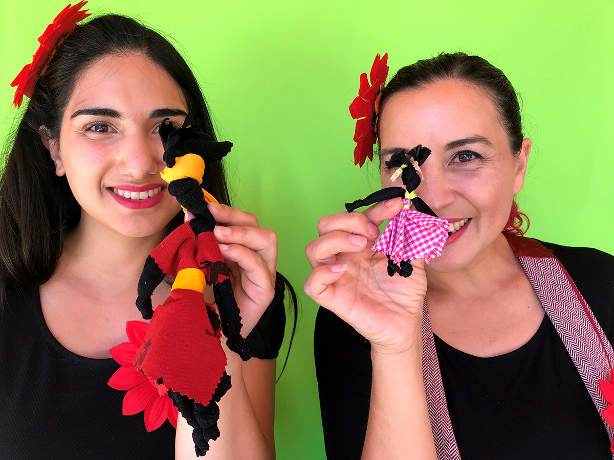 Dos mujeres sonrientes sostienen pequeñas muñecas de tela hechas a mano, vestidas con trajes coloridos, frente a un fondo verde intenso, en una imagen luminosa y festiva.
