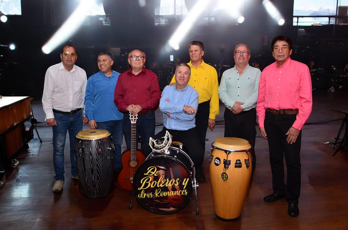 Grupo de músicos adultos posando en un escenario con instrumentos como congas, guitarra y batería con el nombre “De Boleros y otros Romances”, bajo luces teatrales.