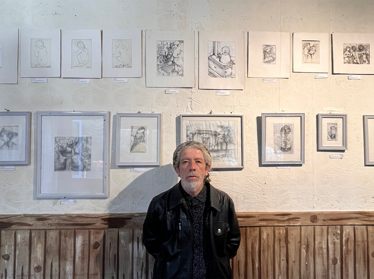 Hombre de pie frente a una pared exhibiendo una colección de dibujos enmarcados en una galería, con fondo de paneles de madera y luz interior cálida. GASTON TOBAR