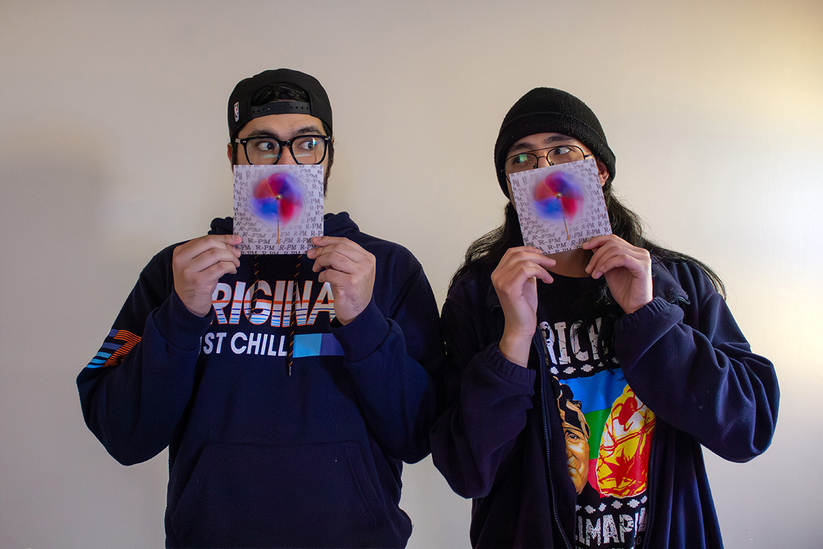 Dos jóvenes sostienen un CD frente a sus rostros, parcialmente cubriéndose la cara, de pie contra una pared blanca. Ambos usan gorro y ropa oscura casual.