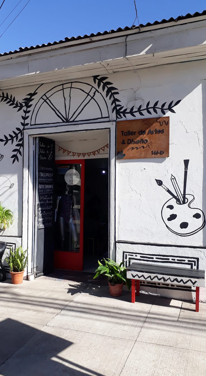 Fachada blanca de un taller de arte y diseño con ilustraciones pintadas en el muro, puerta roja abierta, letrero de madera y plantas en maceteros al exterior.