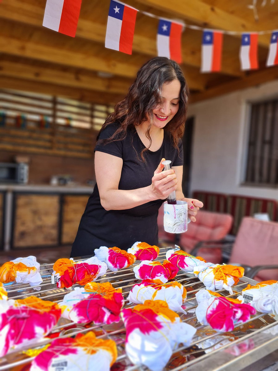 Mujer aplicando tinte con una jeringa sobre telas amarradas con elásticos en una mesa, bajo banderines de Chile.
