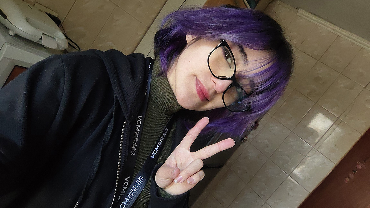Fotografía de una joven con cabello corto morado y lentes, haciendo el gesto de paz con la mano dentro de una sala con piso de cerámica.