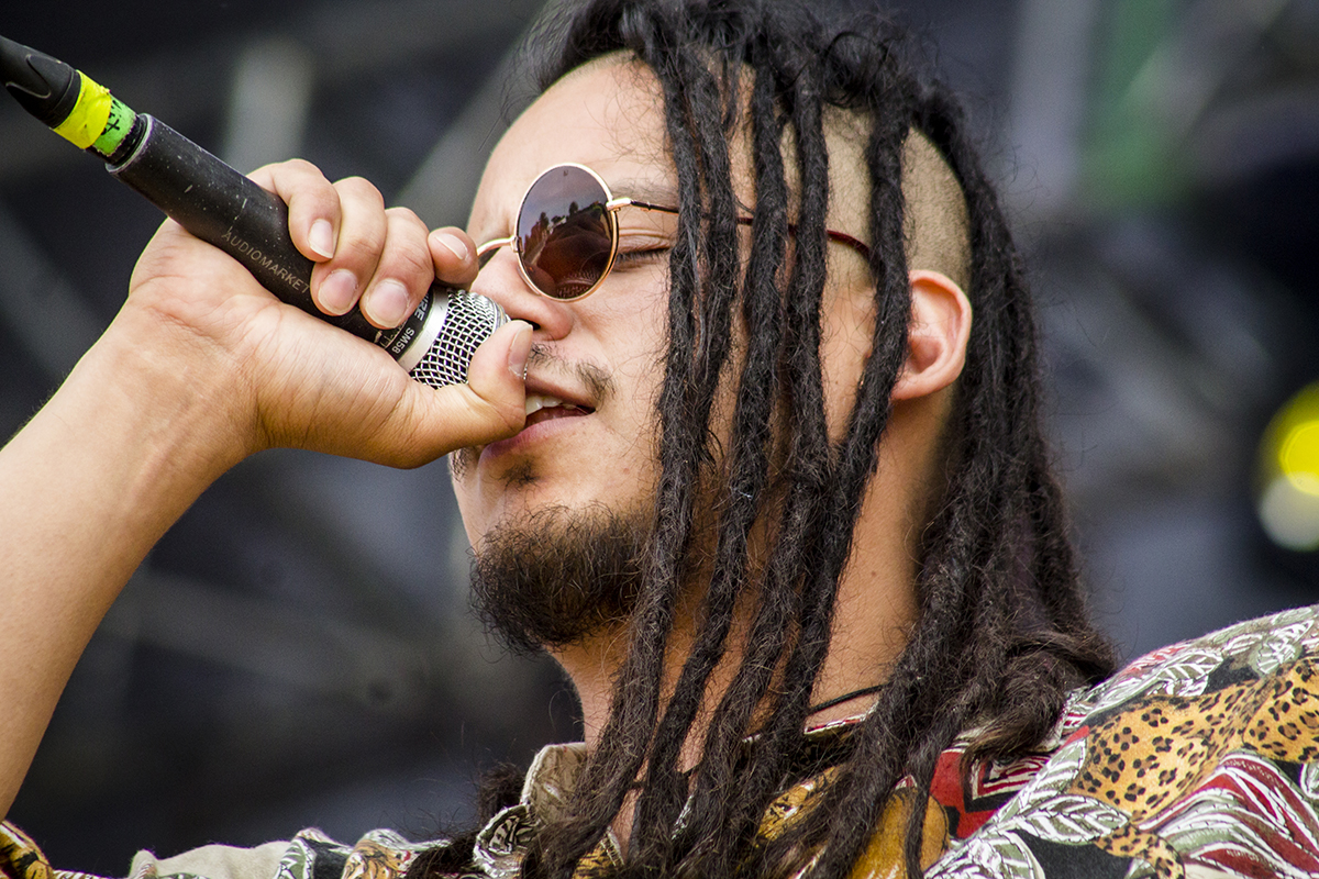 Primer plano de cantante con rastas y gafas oscuras interpretando en vivo con micrófono.