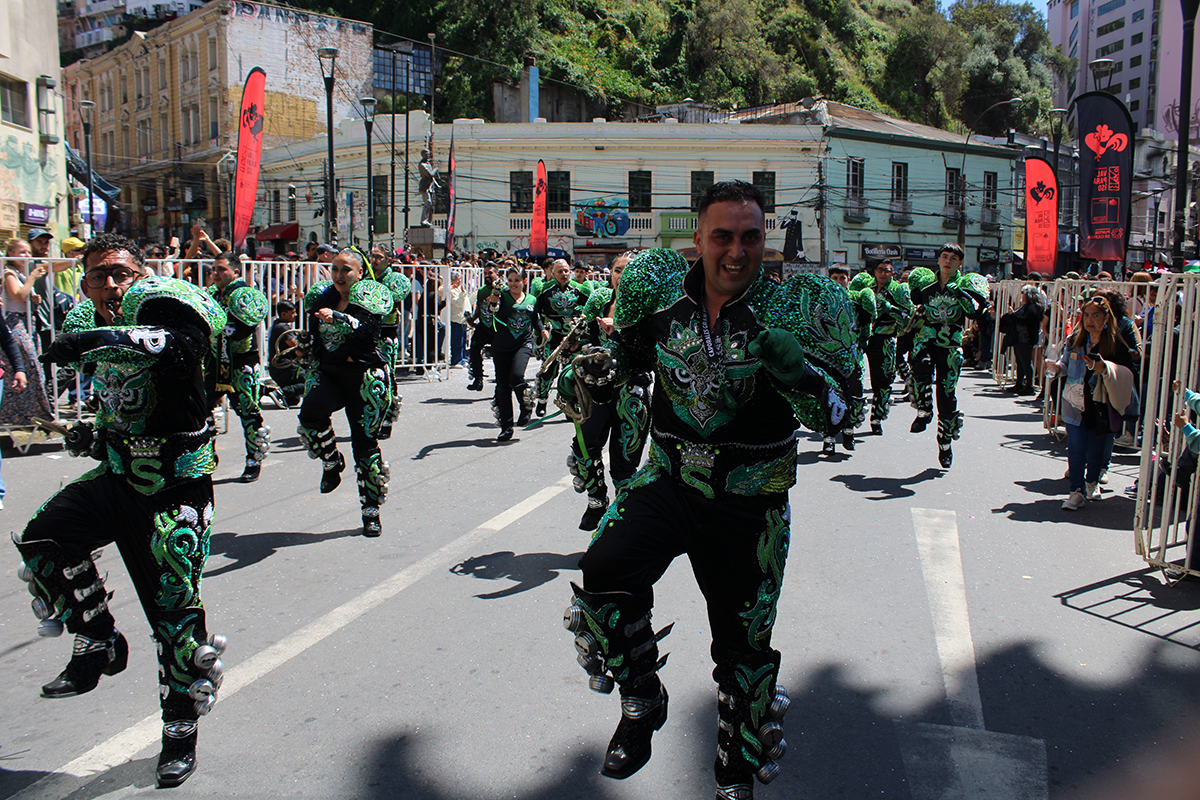 Comparsa de bailarines avanzando por una calle durante un pasacalle urbano, con trajes verdes y negros brillantes y público observando desde las veredas.