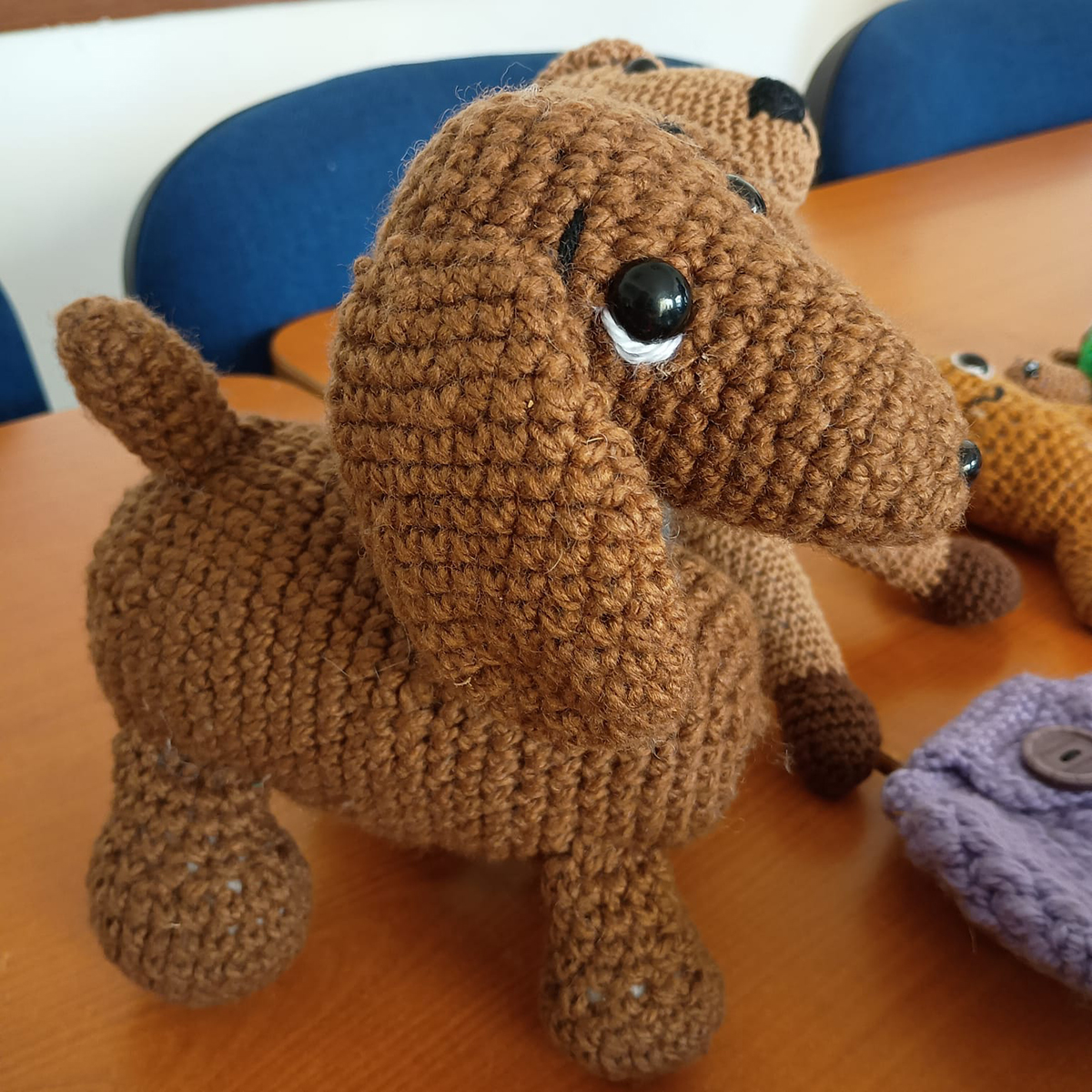 Muñeco tejido a crochet con forma de perro marrón, ojos negros brillantes y orejas largas, sobre una mesa de madera.