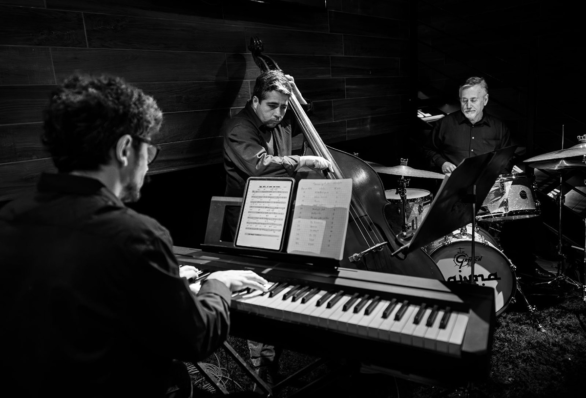 Trío de jazz en blanco y negro interpretando en un espacio interior; pianista leyendo partituras, contrabajista y baterista tocando en ambiente íntimo.
