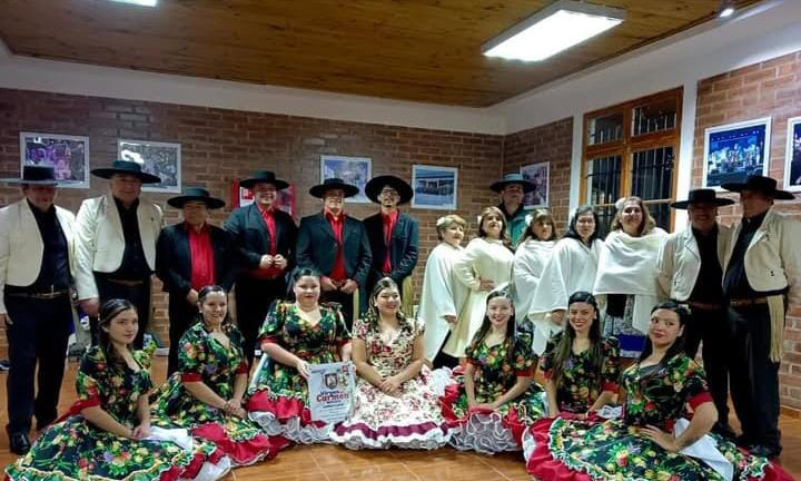 Agrupación folclórica posa en un salón interior con paredes de ladrillo, mujeres con vestidos tradicionales y hombres con sombreros y chaquetas típicas chilenas.