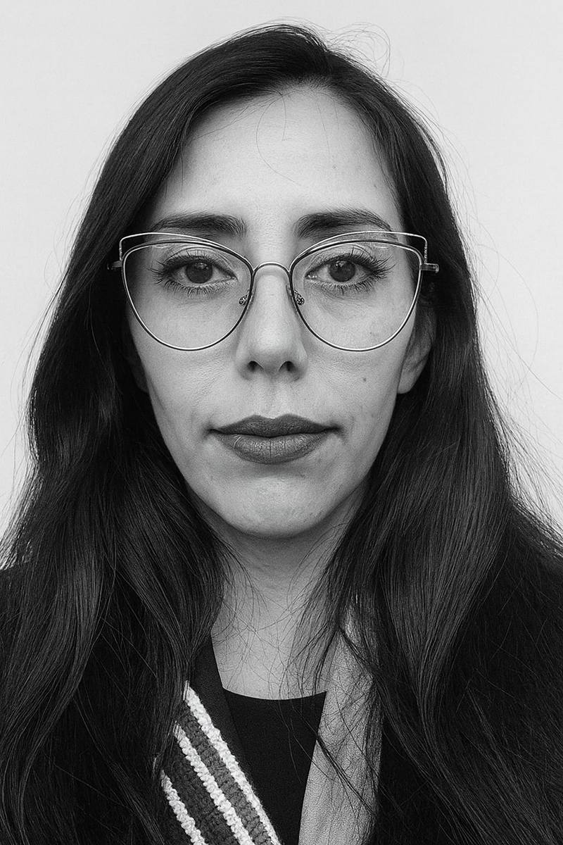 Fotografía en blanco y negro de una mujer de frente, con cabello largo oscuro y lentes de marco metálico, mirando directamente a la cámara sobre fondo neutro.