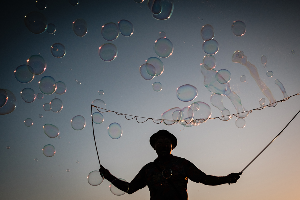 Silueta de un artista callejero sosteniendo varillas para hacer burbujas gigantes contra el cielo al atardecer, rodeado de burbujas flotando.
